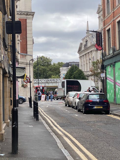 A narrow urban street in Putney during daytime, showing parked cars along the curb, a white moving truck with a closed cargo area positioned at the street corner, and a few people visible near the truck, some carrying or unloading boxes wrapped in cardboard and plastic. The pavement is lined with black bollards to prevent vehicle access, and the adjacent buildings include a shop with signage on the left and a green storefront on the right. In the background, historic-looking white buildings with classical architectural features are visible under a cloudy sky, illustrating the challenges of home relocation and furniture transport along restricted and constrained lanes, as addressed by Putney Removals.