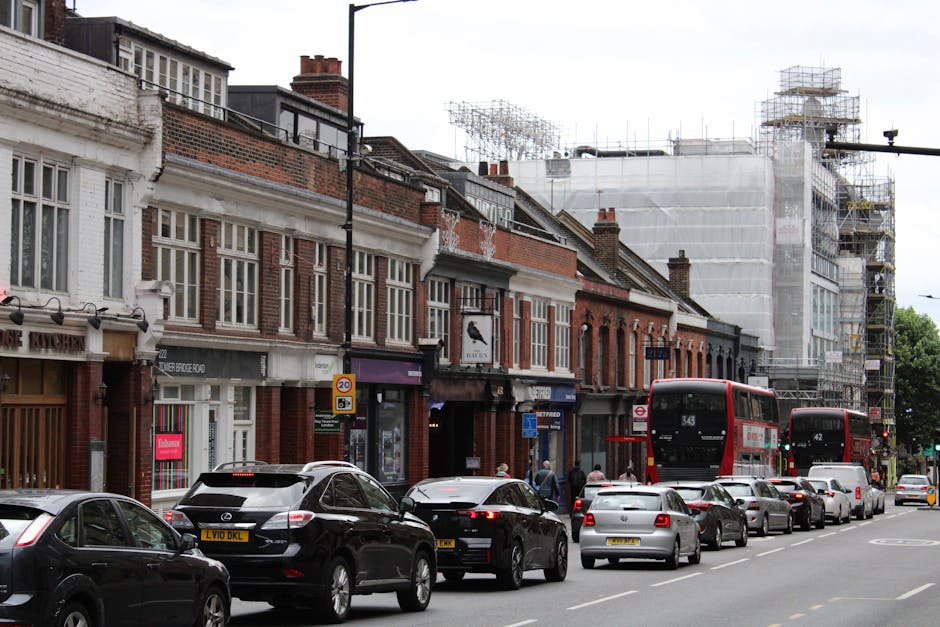 A street scene in Putney showing a row of traditional brick shopfronts and residential buildings, with some upper floors under scaffolding covered by white protective sheeting amid ongoing construction work. Cars, including a black hatchback, a silver sedan, and a white vehicle, are parked along the pavement, while a line of moving and stationary vehicles, such as red double-decker buses and other cars, are visible on the road. In the background, scaffolding surrounds a building under renovation, with construction equipment and metal framework visible. The image captures a daytime setting with overcast weather, lighting providing clear visibility of the scene, and pedestrians walking along the sidewalk. The scene illustrates typical urban residential and commercial street activity, relevant to home relocation and furniture transport logistical considerations, as highlighted by Putney Removals’ services for navigating narrow lanes and access challenges during removals.