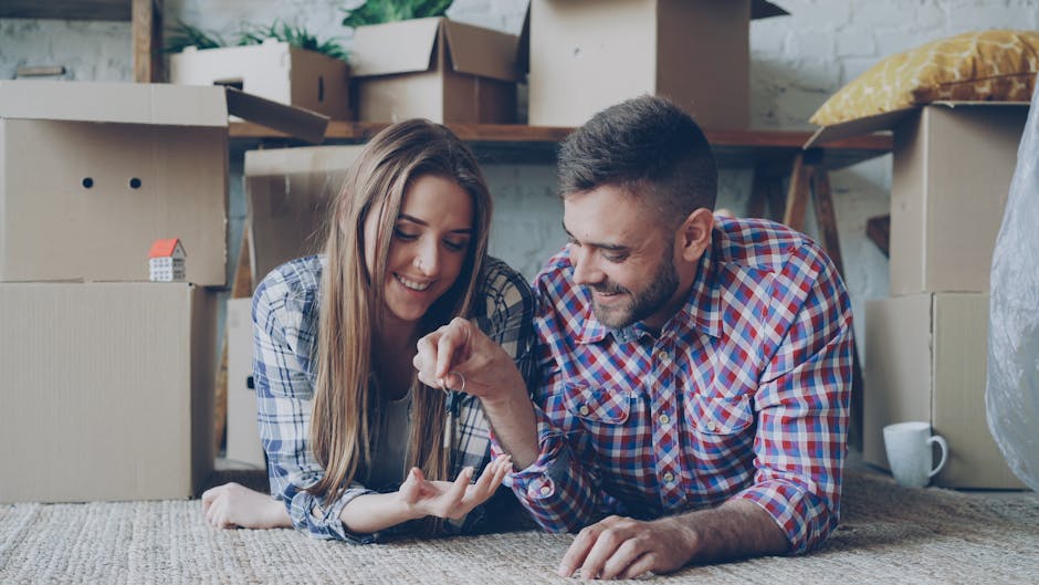 A man and a woman lie on the floor inside a residential property surrounded by packed cardboard boxes, some sealed with packing tape and others open, revealing household items. The woman has long brown hair and wears a blue and white checked shirt, while the man has short dark hair, a beard, and wears a red and blue checked shirt. They are smiling and appear to be engaging in a lighthearted moment during a home relocation process. In the background, there are additional cardboard boxes stacked on shelving and on the floor, some containing small household objects, alongside a few wrapped items. The setting indicates active packing and moving preparations, with the sanded wooden floor and visible door frame suggesting an interior space awaiting furniture transport by [COMPANY_NAME], a professional removals service, as part of the furniture transport and packing process involved in a house move, such as from the Alton Estate to Putney Exchange. The environment is well-lit, with natural light filtering into the room.
