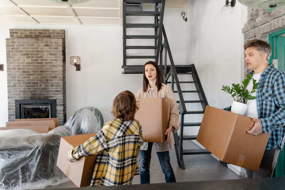 Inside a modern home during a house removal process, two individuals are seen carrying cardboard boxes; a woman with shoulder-length brown hair wearing a beige jacket holds a medium-sized box, while a man with short gray hair wearing a blue checkered shirt carries a larger box with a potted plant inside. A third person, with short brown hair in a yellow and black plaid shirt, is handing over a box to the woman. The background features a contemporary interior with a brick wall, a modern fireplace, and a sleek black staircase leading upstairs. Packaging materials, including plastic-wrapped furniture and additional cardboard boxes, are visible on the floor, indicating an ongoing moving or packing activity. The scene takes place in a well-lit space near a doorway, with household items prepared for transport as part of a furniture transport or home relocation service conducted by Putney Removals, supporting efficient packing and moving logistics.