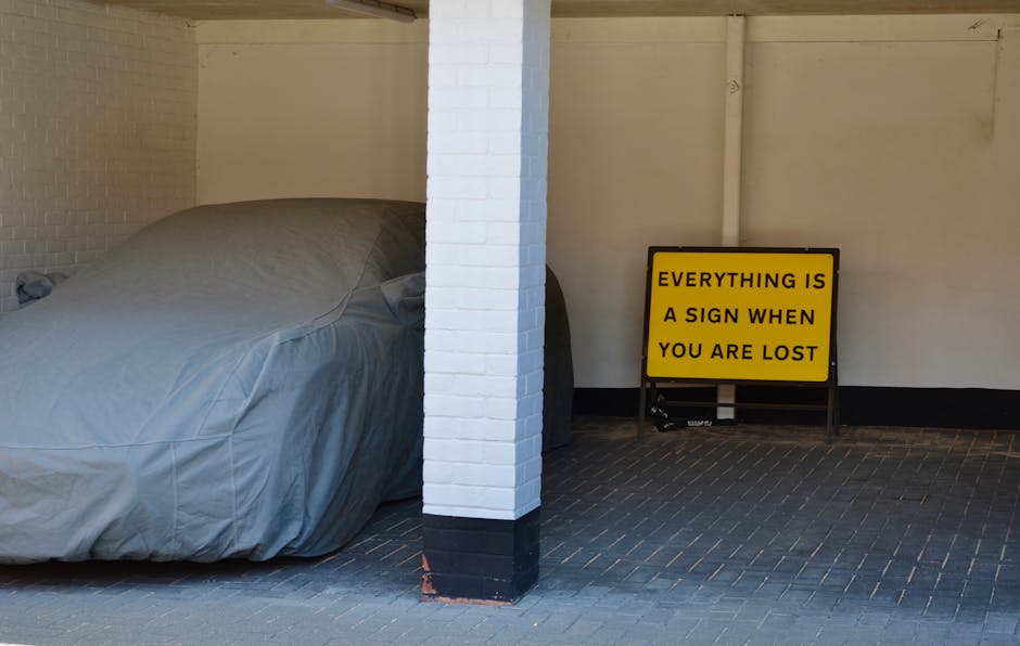 A covered vehicle, possibly a car or small van, parked indoors next to a white-painted brick pillar, with a yellow sign on a stand nearby that reads 'EVERYTHING IS A SIGN WHEN YOU ARE LOST.' The vehicle is draped with a grey protective cover, and the scene appears to be inside a garage or storage area, with a black tiled floor and white walls. The setting includes equipment for home relocation or furniture transport, such as blankets and possibly trolleys, suggesting packing and moving activities guided by Putney Removals. The lighting is natural or ambient, illuminating the covered vehicle and the sign, which emphasizes a helpful message related to moving or navigating during a house relocation process.