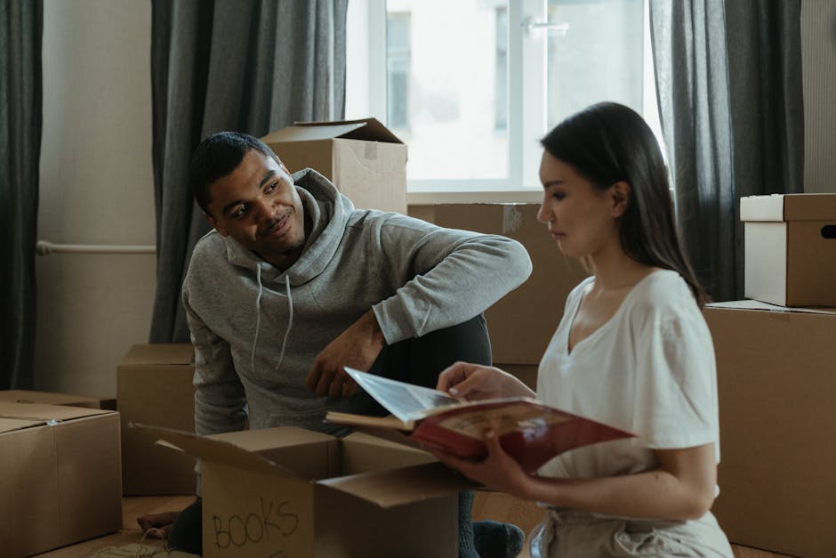 Inside a residential property during a home relocation, a young man with short dark hair, wearing a grey hoodie, is crouched down amidst several packed cardboard boxes, some labeled with items such as 'books.' A young woman with long dark hair, dressed in a white top, is standing nearby holding an open folder or booklet. The boxes are stacked on the wooden floor, with additional boxes and packing materials visible in the background. Natural daylight streams through a window with partially drawn dark curtains, illuminating the room. The scene depicts the packing stage of furniture transport and moving logistics, which could be coordinated by a professional removals service like Putney Removals, supporting careful packing and loading processes involved in house removals.