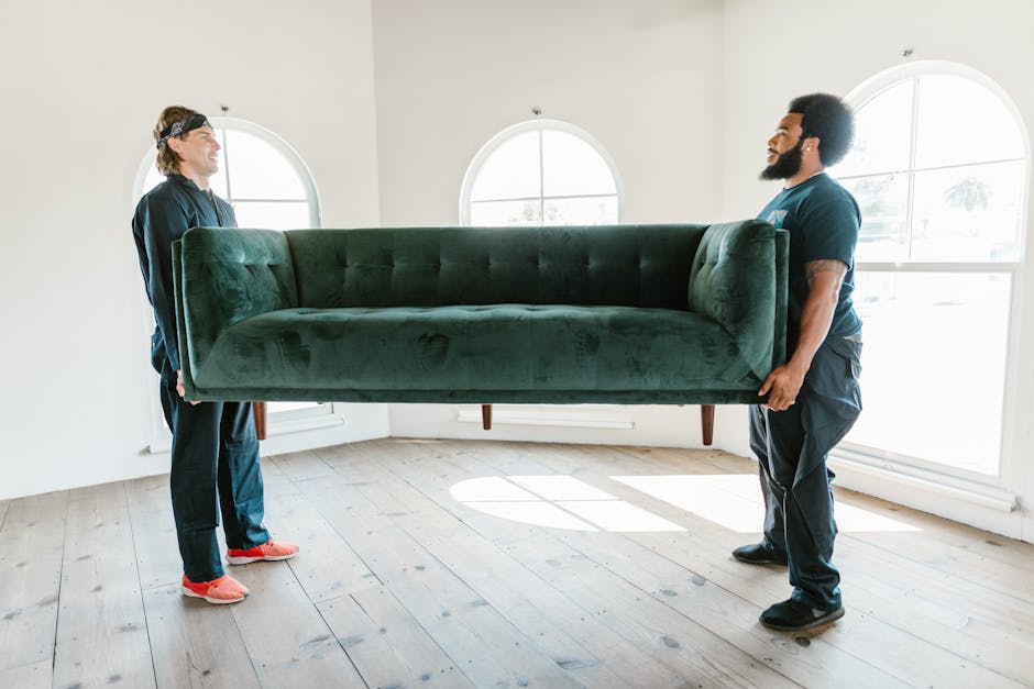 Two movers from Putney Removals are carefully carrying a large, dark green velvet sofa through a spacious, well-lit room with light wooden flooring. The sofa appears to be a three-seater with a tufted backrest and wooden legs. The mover on the left, wearing a navy uniform and pink slippers, is gripping the left armrest, while the mover on the right, also in a navy uniform, is holding the right armrest of the sofa, ensuring stability during the home relocation process. The room features white walls, three large arched windows allowing natural light to flood in, and minimal furnishings. This image depicts part of the furniture transport phase of packing and moving, showcasing careful handling and planning involved in house removals, with the sofa being prepared for loading into a vehicle for transportation to a new property.