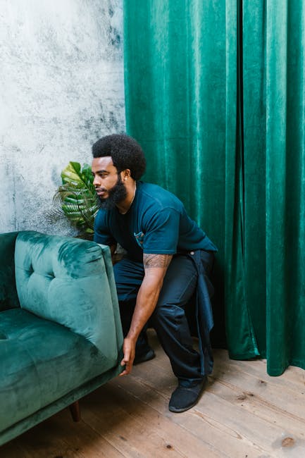 A professional mover from Putney Removals is in the process of preparing a green upholstered sofa for home relocation inside a property. The mover, dressed in a navy blue uniform, is crouched down, carefully lifting the armrest of the sofa. Behind him, there is a textured grey wall and a large indoor plant with broad leaves, situated next to a green curtain that covers part of the window or doorway. The wooden floor is visible, and the lighting is natural, highlighting the detailed fabric of the sofa and the equipment used for furniture transport. The scene captures the meticulous handling involved in loading bulky items like sofas, wardrobes, or pianos onto a moving vehicle, reflecting the logistical aspects of packing and furniture transport services offered by Putney Removals.