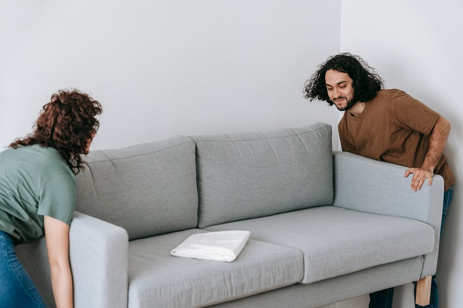 Two movers from Putney Removals are carefully carrying a large, dark green velvet sofa through a spacious, well-lit room with light wooden flooring. The sofa appears to be a three-seater with a tufted backrest and wooden legs. The mover on the left, wearing a navy uniform and pink slippers, is gripping the left armrest, while the mover on the right, also in a navy uniform, is holding the right armrest of the sofa, ensuring stability during the home relocation process. The room features white walls, three large arched windows allowing natural light to flood in, and minimal furnishings. This image depicts part of the furniture transport phase of packing and moving, showcasing careful handling and planning involved in house removals, with the sofa being prepared for loading into a vehicle for transportation to a new property.