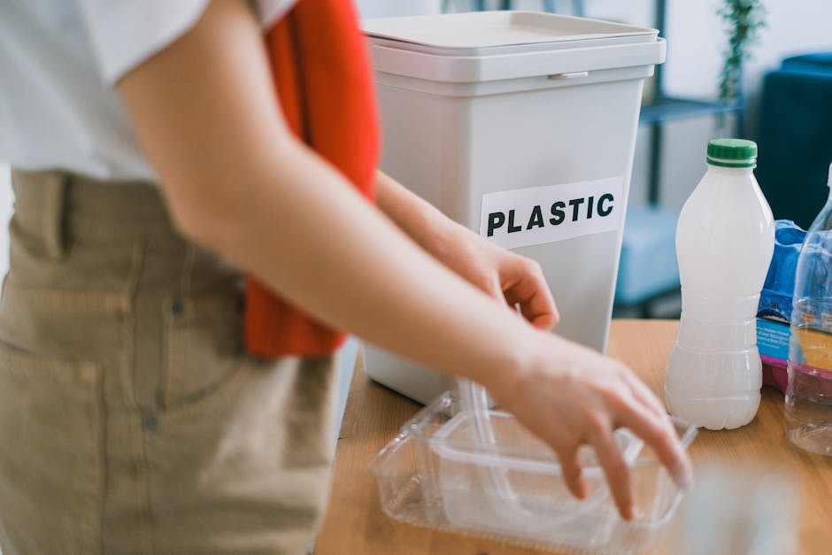 A person wearing a red shirt and beige pants is seen during a home relocation process, placing a clear plastic container with a lid next to a white plastic recycling bin labeled 'PLASTIC,' inside a room with natural lighting. The recycling bin is positioned on a wooden table or counter, with other recyclable items nearby, including a plastic bottle, a jar, and additional containers. The scene captures the packing and sorting aspects of a furniture transport and packing process, emphasizing the separation of plastic waste for disposal or recycling, which is relevant to household waste management and legal disposal rules. In the background, part of a living space with a blue sofa and a potted plant on a side table is visible, indicating the activity is taking place in a residential setting. This visual illustrates aspects of household waste sorting as part of a house removal or moving service, with Putney Removals supporting efficient and compliant packing and disposal procedures after a home move.