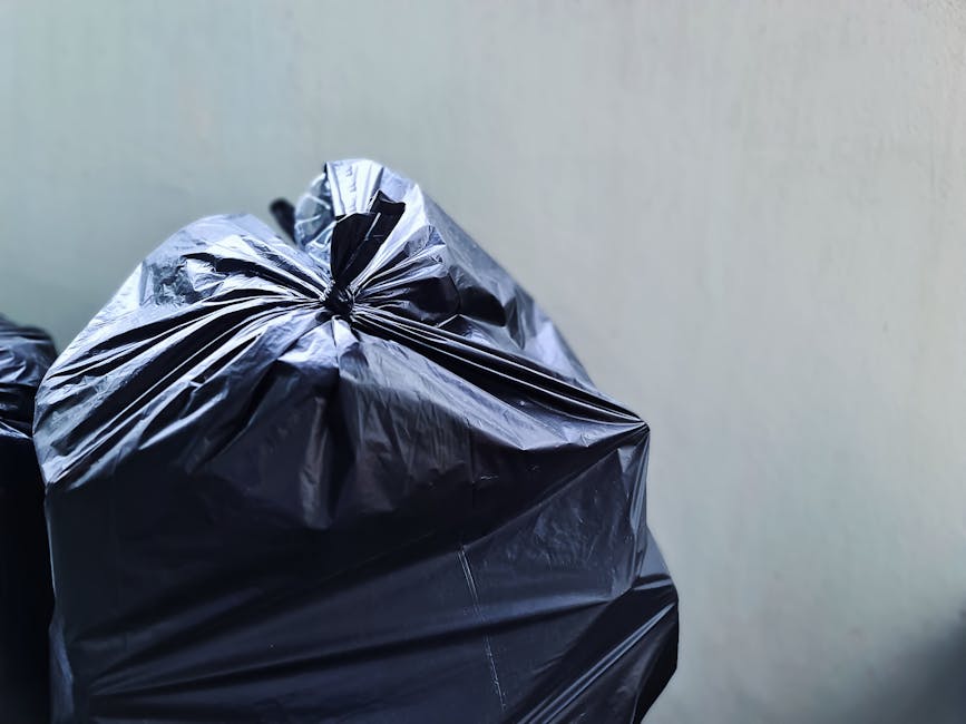 A close-up view of a black trash bag filled with household waste, securely tied at the top, placed against a plain, light-colored interior wall. The bag appears to be made of durable plastic material, commonly used during house relocation or packing to dispose of unwanted items or packing materials such as cardboard, fabric, or plastic wrap. In the background, additional waste bags, also tied and filled, are visible, suggesting a preparation process for waste disposal or furniture transport in a home moving context. The setting is indoors, possibly in a hallway or a room, with a focus on the clutter of waste bags associated with house removals. This scene highlights the importance of proper waste disposal following local regulations as part of a professional home relocation or furniture transport service offered by Putney Removals.
