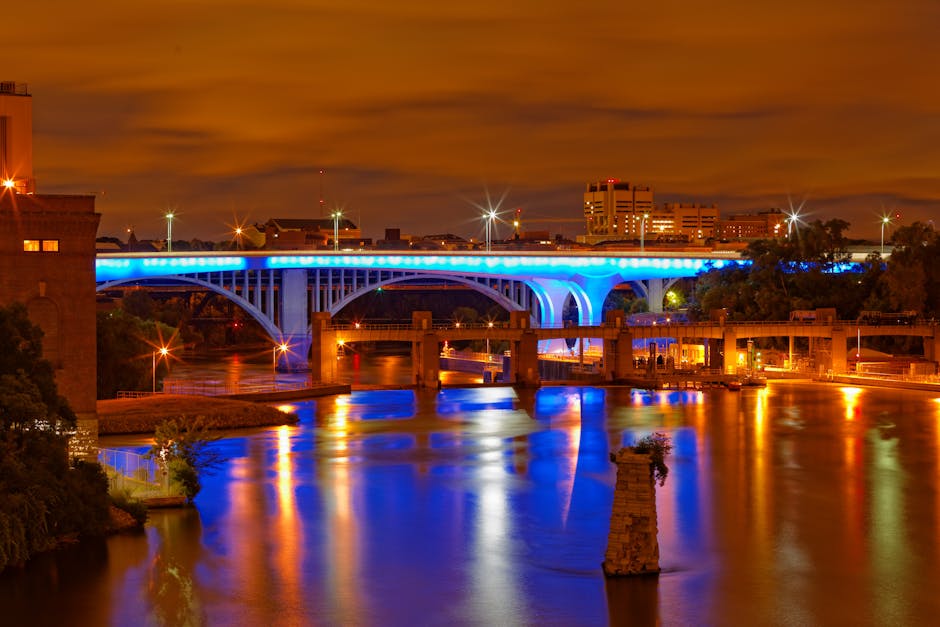 A nighttime view of a city bridge illuminated with blue and white lights, spanning over a calm river that reflects the colorful lighting. The bridge features arched supports and is lined with streetlights that cast bright spots of illumination on the structure. In the foreground, part of a property with a doorway and stone steps is visible, along with several moving cardboard boxes and furniture wrapped in protective plastic and blankets, suggesting active packing or furniture transport. A metal trolley and moving straps are positioned nearby on the pavement, indicating an ongoing loading or unloading process associated with home relocation or furniture transport. In the background, modern buildings and trees are illuminated by streetlights, under a cloudy evening sky with an orange glow. This scene is related to house removals, featuring logistics and packing elements that align with the context of moving near Putney Bridge, as provided by Putney Removals.