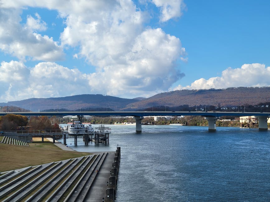 A nighttime view of a city bridge illuminated with blue and white lights, spanning over a calm river that reflects the colorful lighting. The bridge features arched supports and is lined with streetlights that cast bright spots of illumination on the structure. In the foreground, part of a property with a doorway and stone steps is visible, along with several moving cardboard boxes and furniture wrapped in protective plastic and blankets, suggesting active packing or furniture transport. A metal trolley and moving straps are positioned nearby on the pavement, indicating an ongoing loading or unloading process associated with home relocation or furniture transport. In the background, modern buildings and trees are illuminated by streetlights, under a cloudy evening sky with an orange glow. This scene is related to house removals, featuring logistics and packing elements that align with the context of moving near Putney Bridge, as provided by Putney Removals.