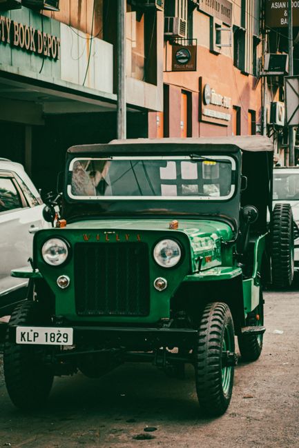 A vintage green utility vehicle with a boxy front and round headlights is parked on a street in an urban area. The vehicle features a black grille and windshield, with a metal roof supported by visible frame structures. Behind the vehicle, a modern white car is partially visible. The background includes multi-storey buildings with a mix of commercial signage, including a bookstore and various shop signs, and several air conditioning units mounted on the exterior walls. The scene appears to be captured during daytime with natural lighting. The image reflects an environment where home relocation or furniture transport might occur, illustrating vehicle options used in local removals services, such as those offered by Putney Removals. The vehicle is positioned on the pavement, and there are no visible moving activities in progress in this specific shot.