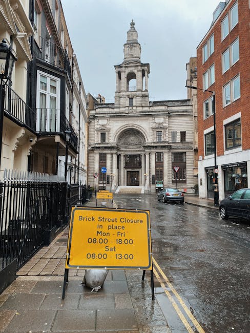 A wet street scene in Putney showing old and modern buildings on either side of the road, with a historic stone church at the end. In the foreground, a yellow road closure sign indicates restrictions for street work and parking, specifying times from Monday to Saturday. The sign is positioned on the pavement in front of a black metal railing and is secured by a weighted base. Several parked vehicles are visible along the street, with one car approaching the open road. The pavement appears slick from recent rain, and grey clouds fill the sky above. This setting illustrates a typical urban environment suitable for home relocation or furniture transport, with ongoing street work affecting vehicle and moving logistics, supported by Putney Removals' professional services.