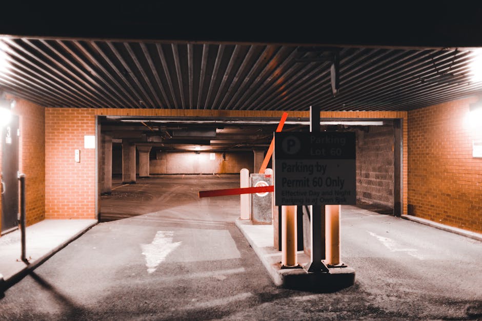 The image shows the entrance to an underground parking garage with a lowered security barrier arm made of orange plastic, positioned across the driveway. In front of the barrier, there is a parking regulation sign mounted on a black post, displaying parking lot information such as lot number 60 and permit requirements. The parking area beyond the barrier is dimly lit with concrete and brick walls, with several support columns visible in the background. On the left side of the entrance, there is a wall with a grey metal door and a small white sign, suggesting access control or building entry. The ceiling of the garage features dark wooden or metal slats running parallel, and the ground is paved with asphalt, marked by a white arrow pointing forward. This setting is indicative of a secure parking facility used during home relocation or furniture transport processes managed by Putney Removals, supporting efficient movement of boxes, furniture, and removal equipment in the context of house removals or moving services.