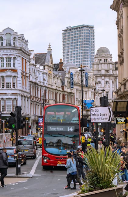 A busy urban street scene in Putney showing a double-decker bus with the route number 14 to Putney Heath parked at a bus stop. Surrounding the bus are several pedestrians on the pavement, some carrying bags or backpacks, and others waiting to cross or board the bus. To the right, there is a small group of people near shop entrances, including a tobacco shop with a yellow sign. In the foreground, a large green plant with spiky leaves is visible on the sidewalk. The background features historic Victorian-style buildings with ornate facades and decorative elements, alongside modern high-rise structures under an overcast sky. Visible street signs include directions to Trafalgar Square and Hyde Park Corner, with street lamps lining the road. The scene captures the typical movement associated with home relocation and furniture transport in central London, with vehicles, packaging materials, and the activity of moving residents contributing to the urban atmosphere. Putney Removals ensures professional logistics for packing, loading, and transport during house moves.